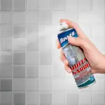 Mano sosteniendo aerosol Binner Quita Sarro 400ml, aplicando espuma blanca sobre baldosas grises en baño. Etiqueta azul y roja visible.