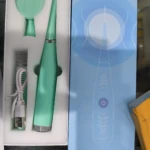 Limpiador eléctrico de sarro en color verde menta, con cepillo de limpieza, base de carga y cable USB, presentado en caja abierta junto a su empaque azul.