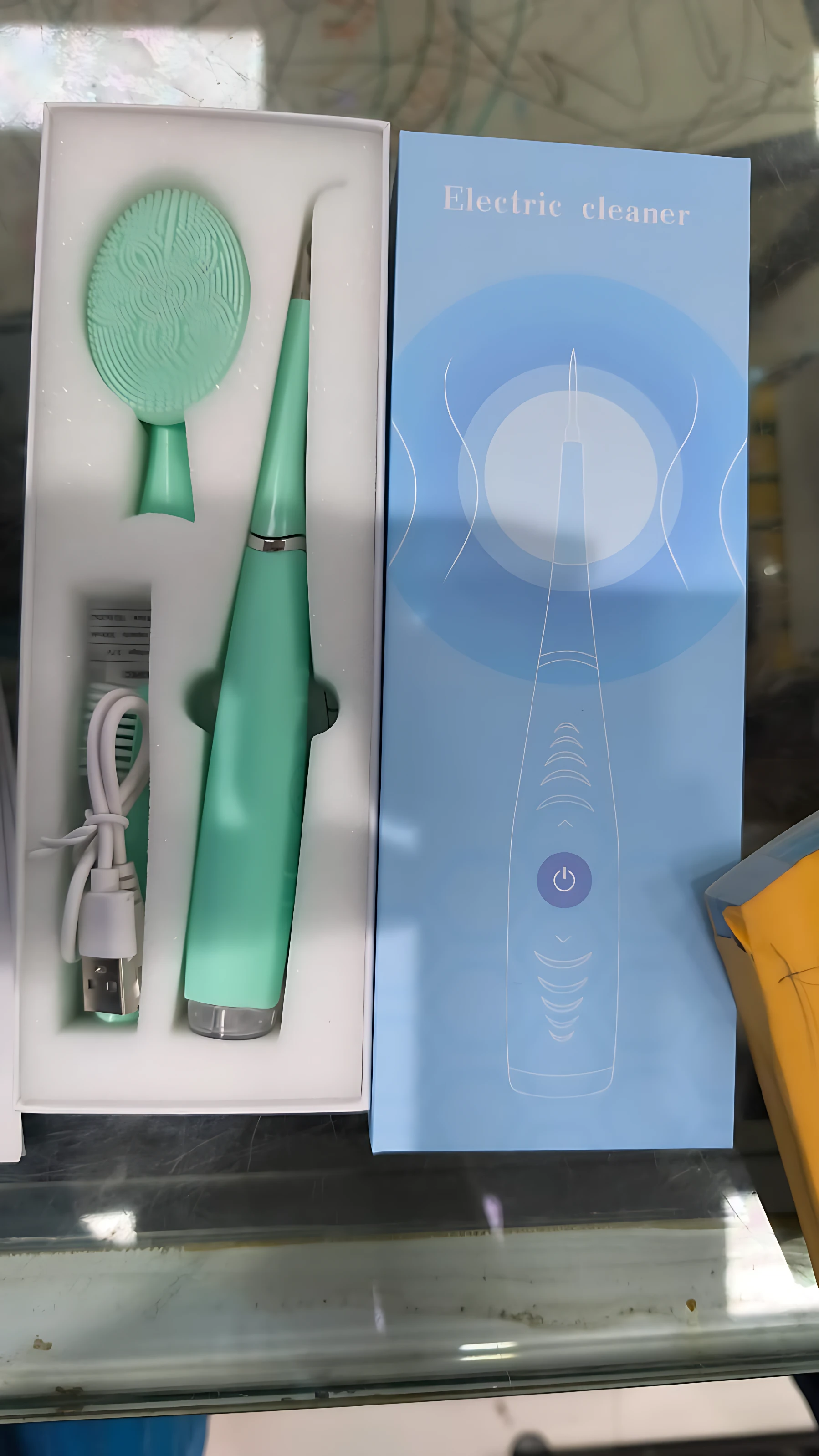 Limpiador eléctrico de sarro en color verde menta, con cepillo de limpieza, base de carga y cable USB, presentado en caja abierta junto a su empaque azul.