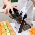 Rallador mandoline blanco con mango negro, en uso para cortar zanahorias y pepino en rodajas finas sobre tabla de madera.