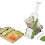 Rallador mandoline verde con mango seguro, mostrando rodajas de pepino, zanahoria y queso sobre tabla de madera, junto a accesorios incluidos.