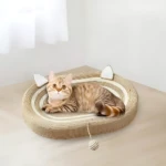 Gato durmiendo en cama rascadora ovalada de fibras naturales con orejas decorativas y juguete colgante, sobre superficie clara.