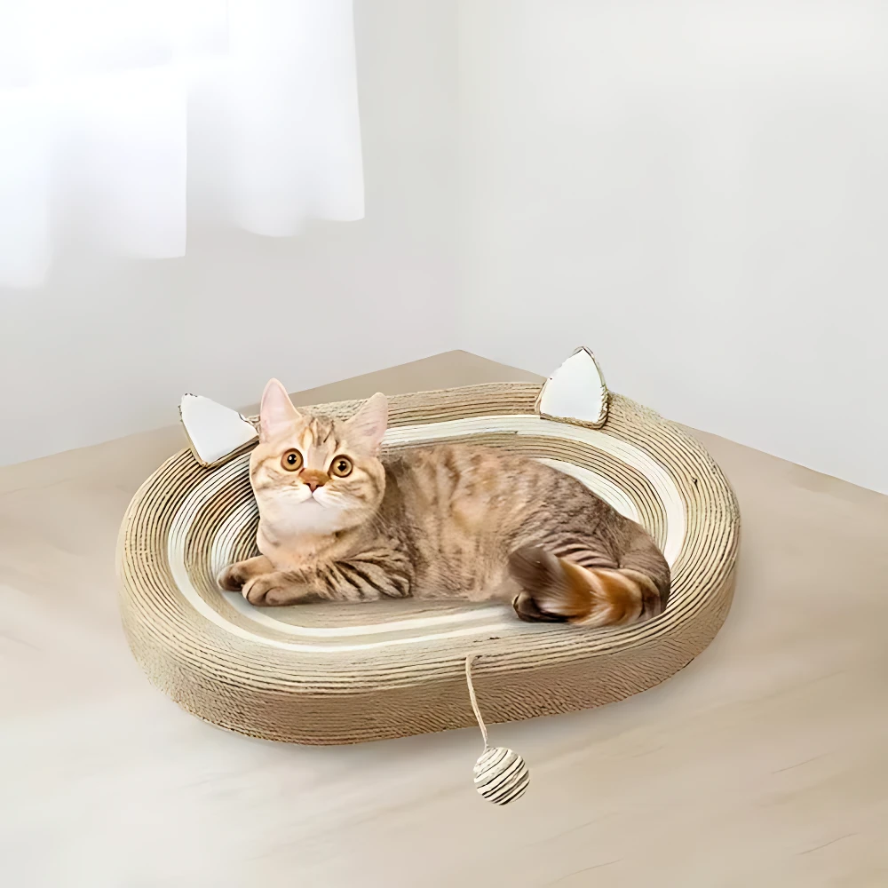 Gato durmiendo en cama rascadora ovalada de fibras naturales con orejas decorativas y juguete colgante, sobre superficie clara.