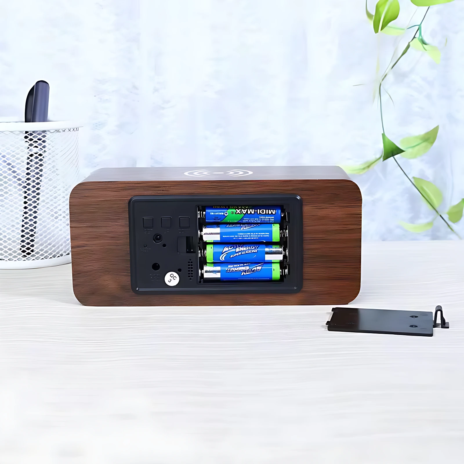Reloj despertador de madera con panel trasero abierto mostrando cuatro pilas AA y puerto de carga inalámbrica, sobre escritorio blanco.