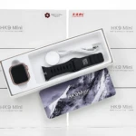 Reloj inteligente HK9 Mini en caja abierta, mostrando pantalla cuadrada, correa negra, cargador y cable. Fondo con múltiples cajas blancas del modelo.