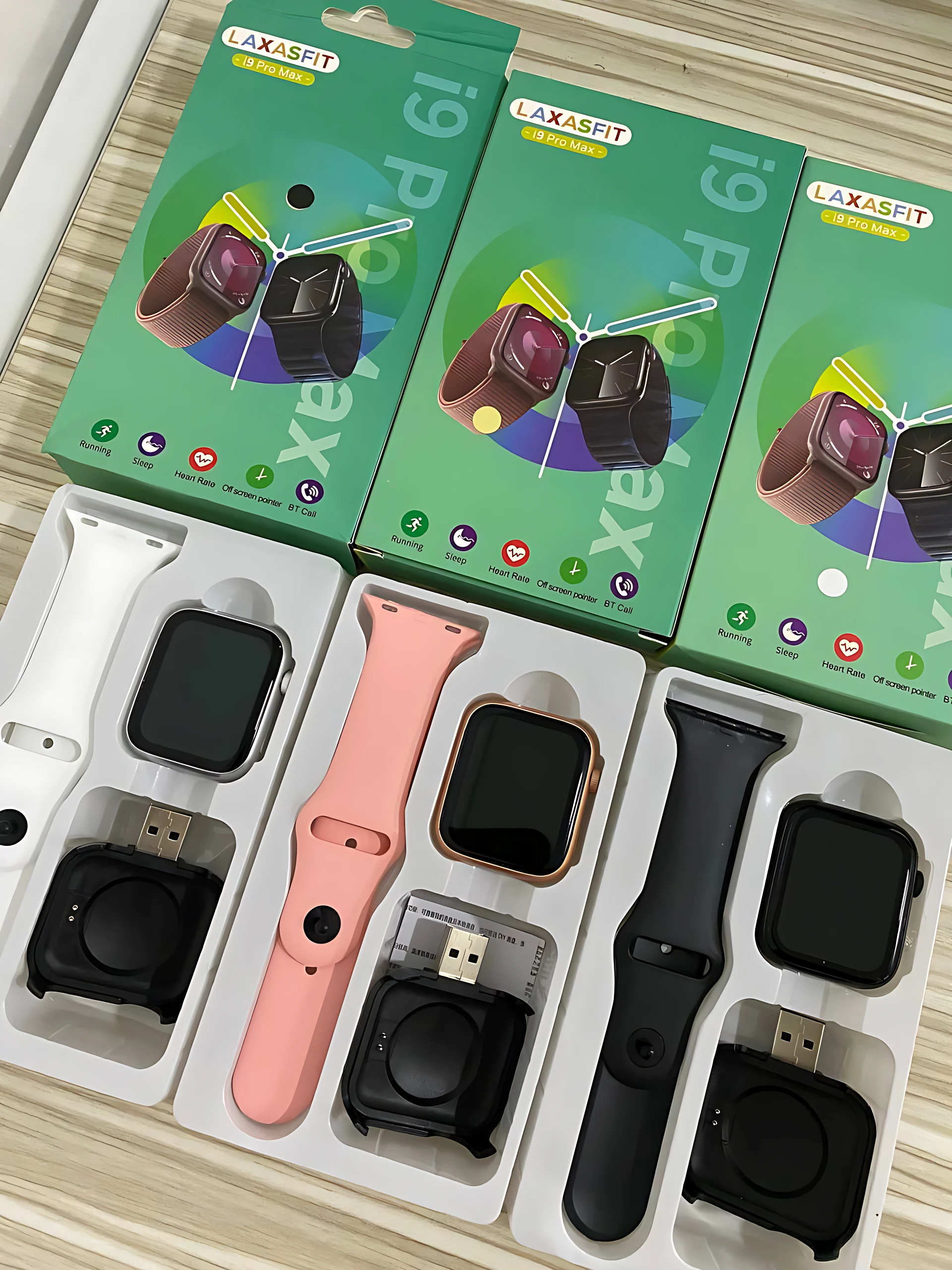 Pack de tres relojes inteligentes I9 Pro Max con correa intercambiable en blanco, rosa y negro, cada uno con cargador magnético incluido, presentados en cajas verdes sobre superficie de madera.