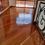 Piso de madera pulido con acabado brillante, reflejando luz natural desde ventana grande; detalles de barandilla metálica y armario empotrado en fondo.