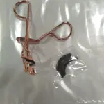 Rizador de pestañas en acero con mango en tono cobre, acompañado de una pieza de repuesto negra, ambos dentro de su empaque transparente.