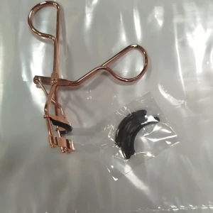 Rizador de pestañas en acero con mango en tono cobre, acompañado de una pieza de repuesto negra, ambos dentro de su empaque transparente.