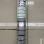 Rollo adhesivo pared acolchado gris con etiquetas informativas, envuelto en plástico transparente, sobre fondo de baldosas beige.