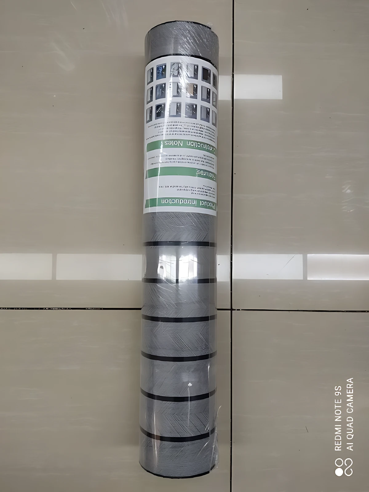 Rollo adhesivo pared acolchado gris con etiquetas informativas, envuelto en plástico transparente, sobre fondo de baldosas beige.