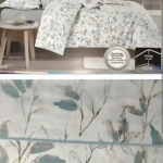 Colcha estampada floral doble faz en tonos claros con hojas verdes, vista frontal en cama con cojines y mesita lateral; incluye etiqueta de calidad premium.
