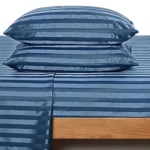 Sábanas de polialgodón con rayas horizontales en tonos azul oscuro y gris, sobre colchón alto con base de madera clara.