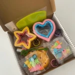 Caja abierta con moldes de acero inoxidable en forma de estrella, corazón y flor, junto a accesorios de colores para decorar galletas.