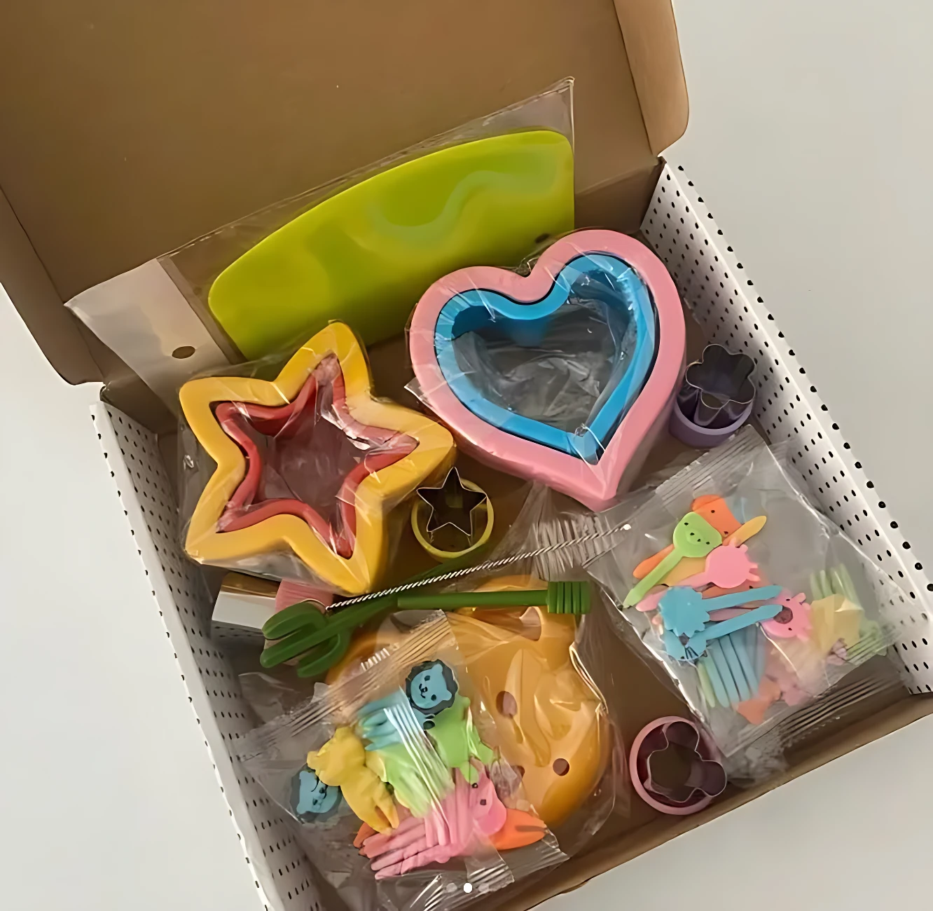 Caja abierta con moldes de acero inoxidable en forma de estrella, corazón y flor, junto a accesorios de colores para decorar galletas.