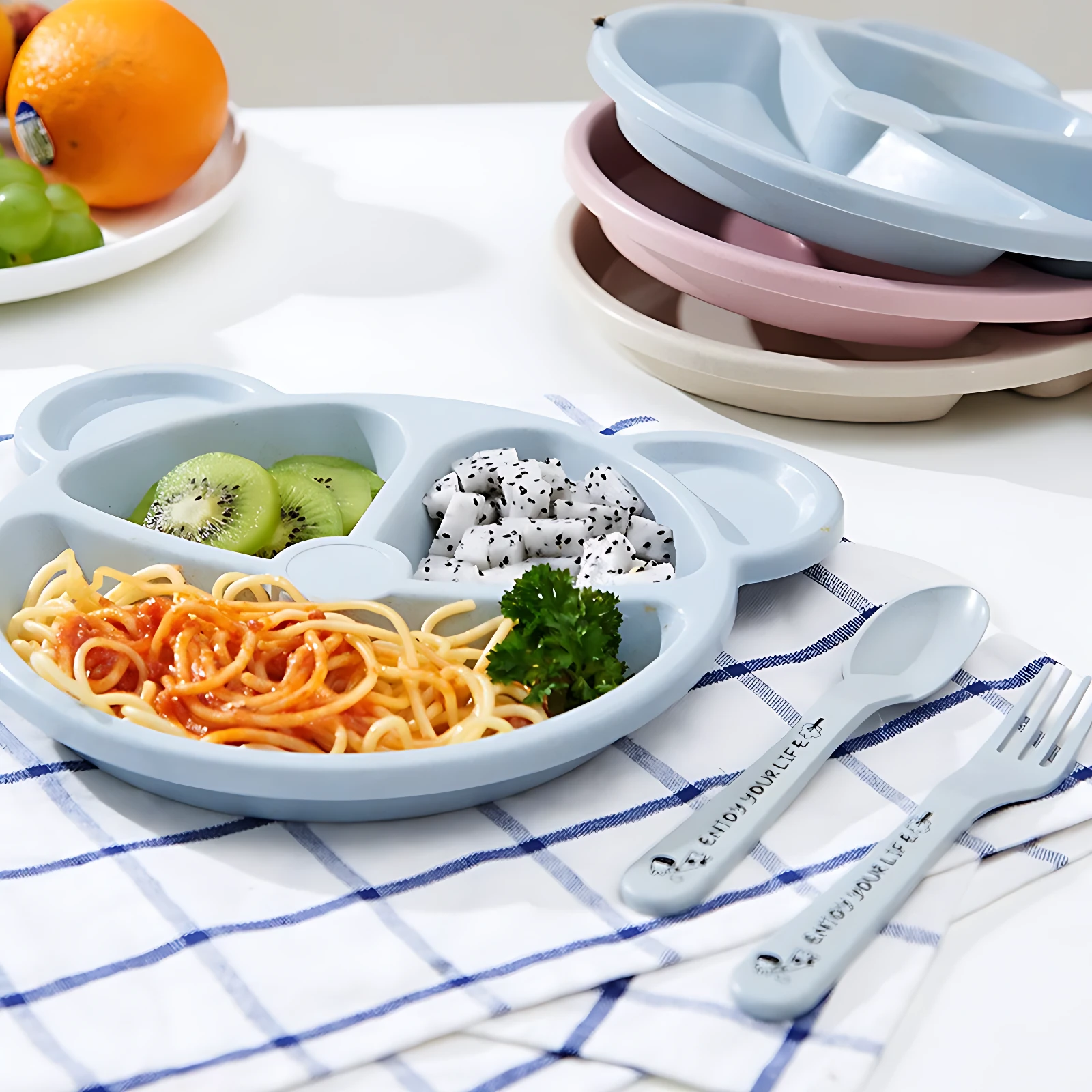 Plato infantil de fibra de trigo en color azul claro con tres compartimentos, mostrando pasta, kiwi y fruta. Incluye cubiertos y vajilla apilada en tonos pastel.