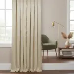 Cortina de poliéster beige con aro metálico, colgada en ventana, con pliegues verticales y caída suave.