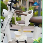 Silla infantil blanca con asiento marrón y arnés, montada en la parte delantera de un scooter eléctrico. Incluye reposabrazos y base ajustable para mayor seguridad.
