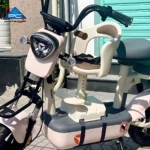 Silla infantil delantera beige con arnés ajustable, montada en scooter eléctrico de color crema, con asiento acolchado y soporte para pies.