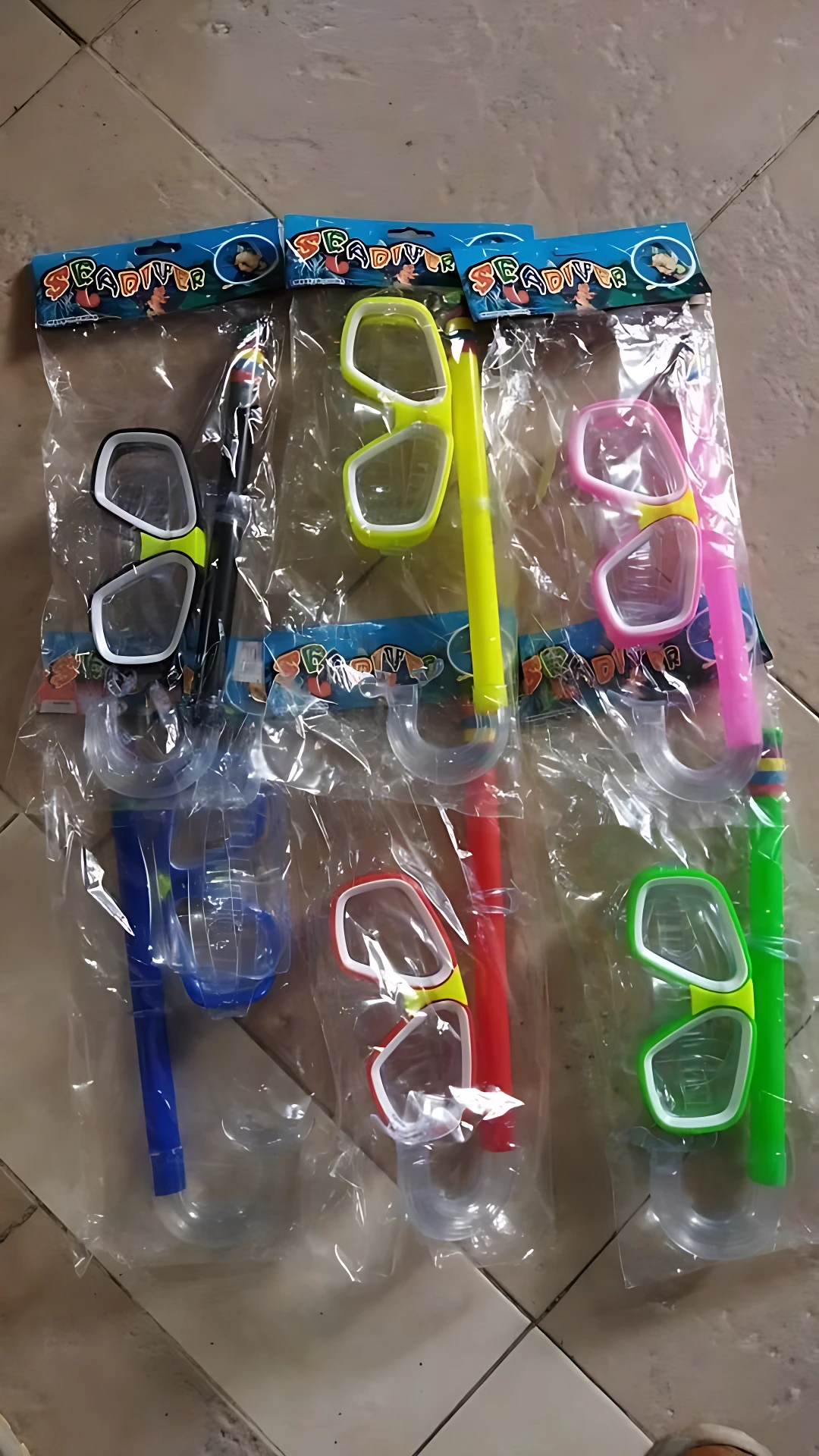 Seis snorkel con caretas de colores variados, incluyendo azul, verde, rosa y amarillo, dispuestos en paquetes individuales sobre un fondo de baldosas.