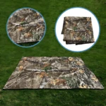 Sobre carpa de camuflaje con 4 extensiones para anclar, desplegado sobre césped verde; incluye dos vistas circulares: una ampliada de textura y otra plegada mostrando capas.