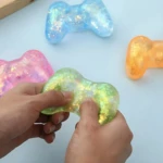 Squishy de control X12 en verde con brillo interior, siendo apretado por dos manos; otras versiones en azul, rosa y naranja visibles en el fondo.