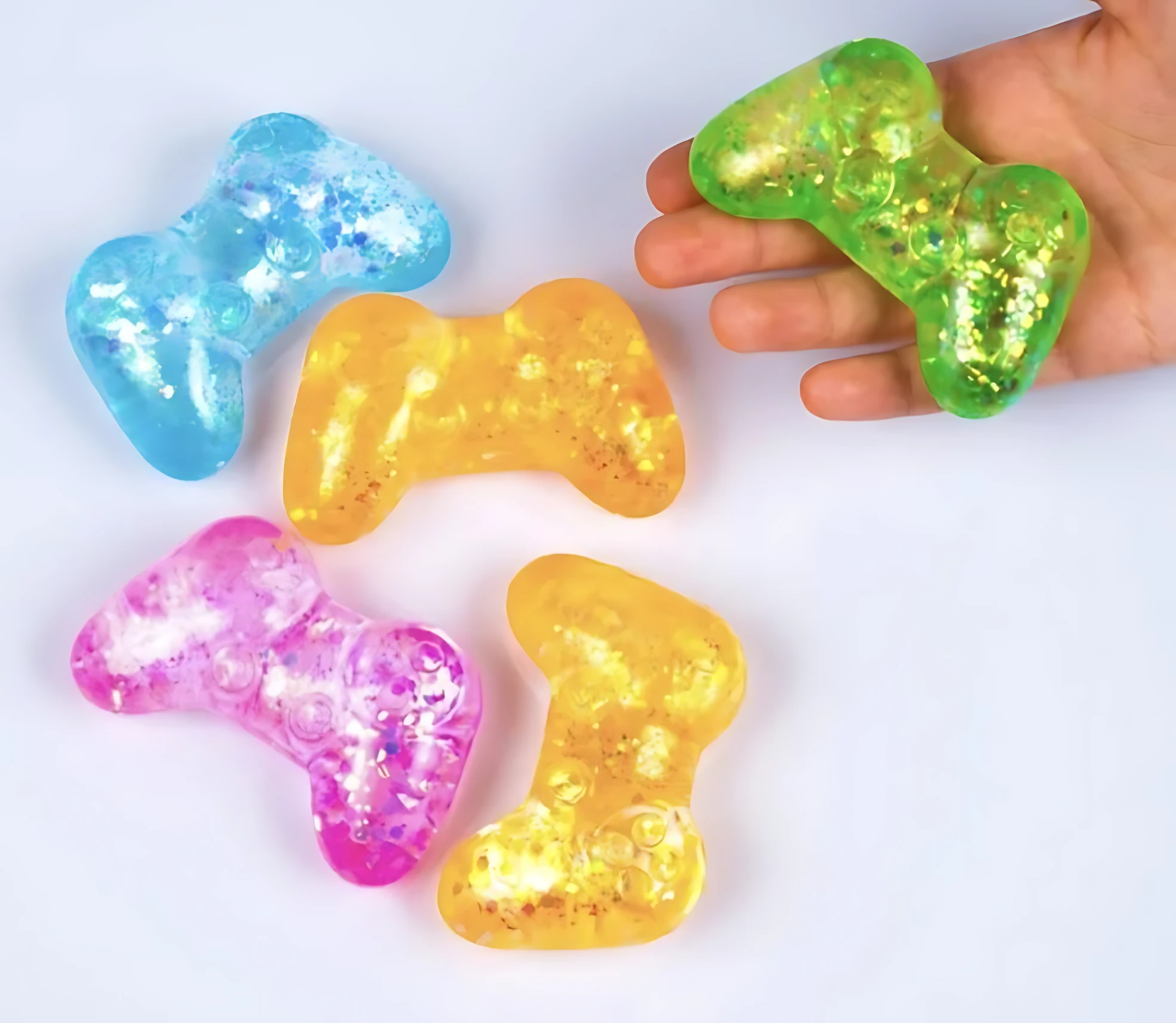 Cinco juguetes antiansiedad en forma de control de videojuegos, colores azul, amarillo, rosa y verde con brillo interior, uno sostenido en mano.