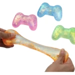 Squishy en forma de control de videojuegos con brillo interior, en tonos naranja, rosa, azul y verde. Se muestra siendo estirado entre manos, destacando su textura elástica y partículas brillantes.
