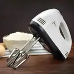 Batidora de mano blanca con accesorios metálicos, sobre mesa de madera, junto a tazón de harina y huevos.
