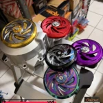 Tapa volante giratorio de aluminio anodizado en colores dorado, rojo, negro, morado y multicolor, montada sobre estructura metálica, ideal para personalizar scooters.