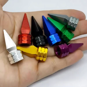 Siete tapas de válvula tipo spike en aluminio pulido, colores rojo, negro, azul, verde, plateado, amarillo y morado, dispuestas sobre mano abierta.