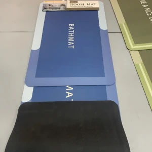 Tapete de baño antideslizante en tonos azul y negro, con borde blanco y etiqueta que indica "ROOM MAT". Se muestra desplegado sobre suelo de baldosas, junto a otra variante en verde oliva.