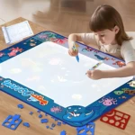 Niña dibujando en tapete mágico de agua con temática oceánica, bordes decorados con animales marinos y piezas de rompecabezas dispersas sobre mesa de madera.