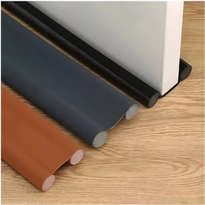 Tira de sellado para puerta en tres colores: negro, gris oscuro y marrón. Vista lateral mostrando perfil redondeado y base adhesiva, sobre superficie de madera clara.