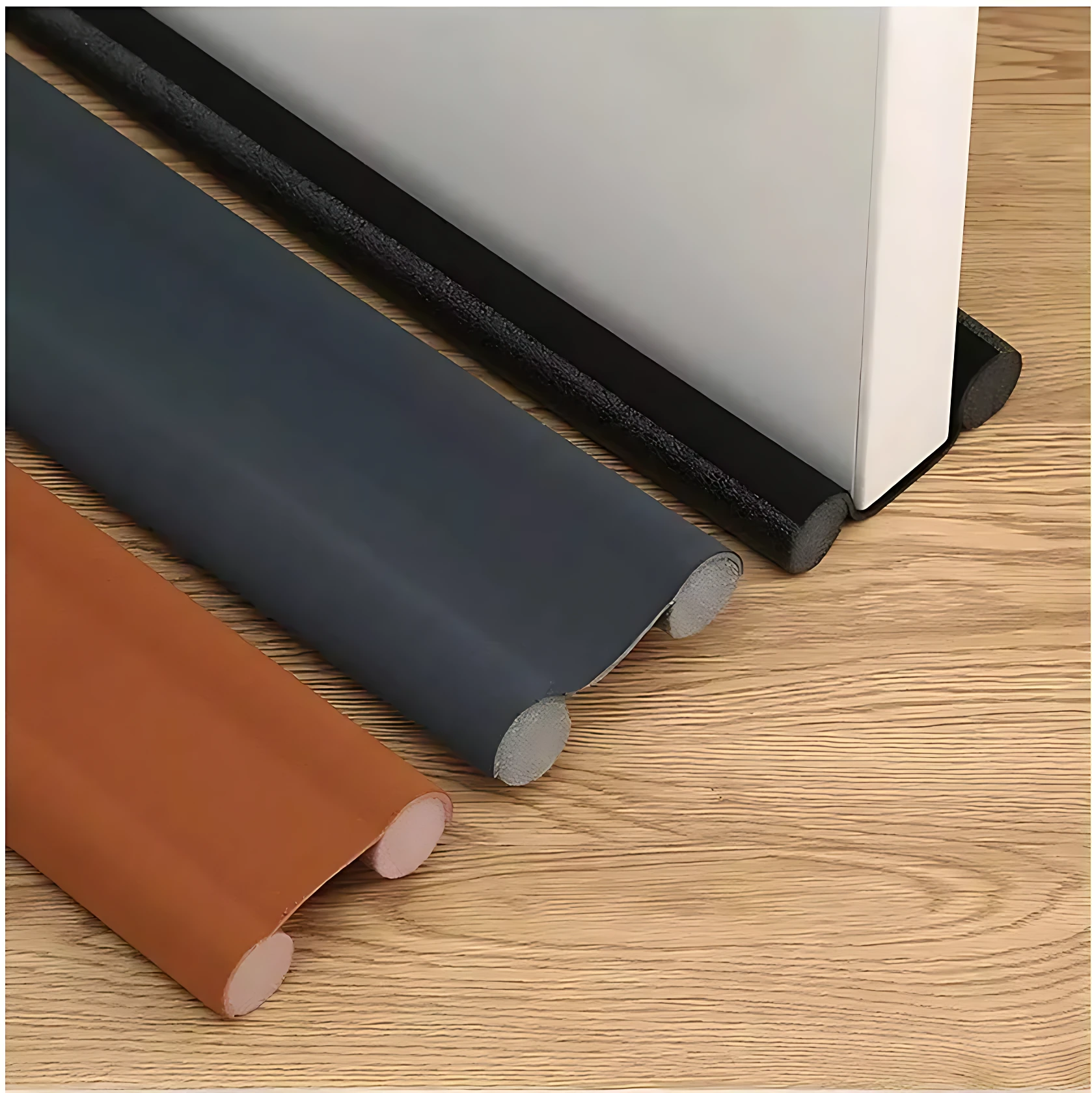Tira de sellado para puerta en tres colores: negro, gris oscuro y marrón. Vista lateral mostrando perfil redondeado y base adhesiva, sobre superficie de madera clara.