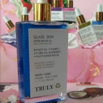 Botella de vidrio azul con tapa dorada y cuentagotas, etiquetada como Glass Skin After Shine Oil de Truly, con ingredientes visibles: Bakuchiol, Vitamina C, Jojoba Oil, Blueberry y Niacinamide Boost. Presentación en set con múltiples unidades y accesorios incluidos.
