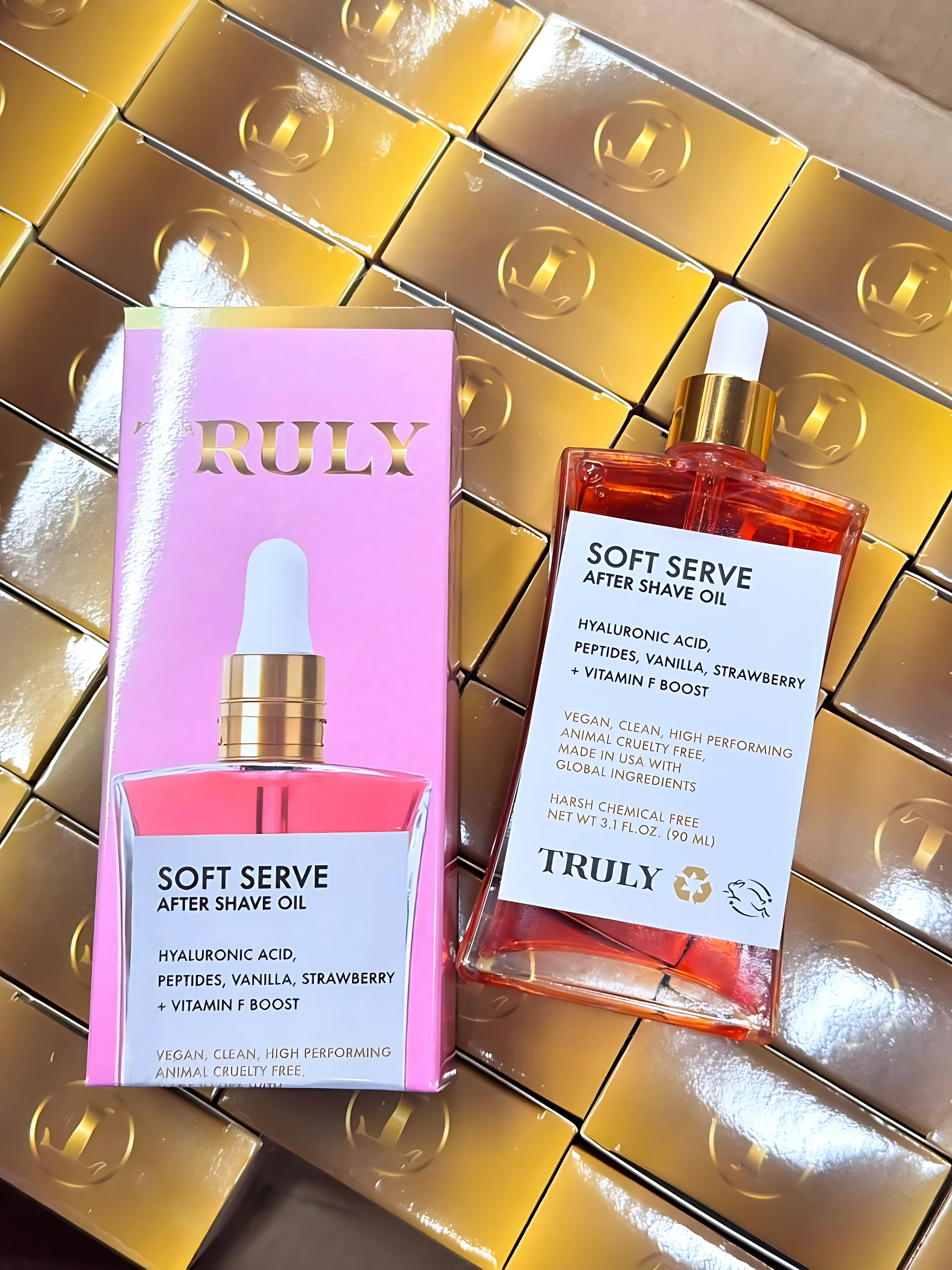 Packaging rosa y frasco de aceite facial Truly Soft Serve After Shave Oil con gotero, sobre cajas doradas.