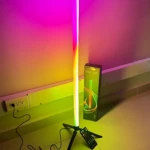 Tubo neón RGB vertical de 75 cm con soporte trípode negro, emitiendo luz multicolor en ambiente interior junto a su caja y control remoto.