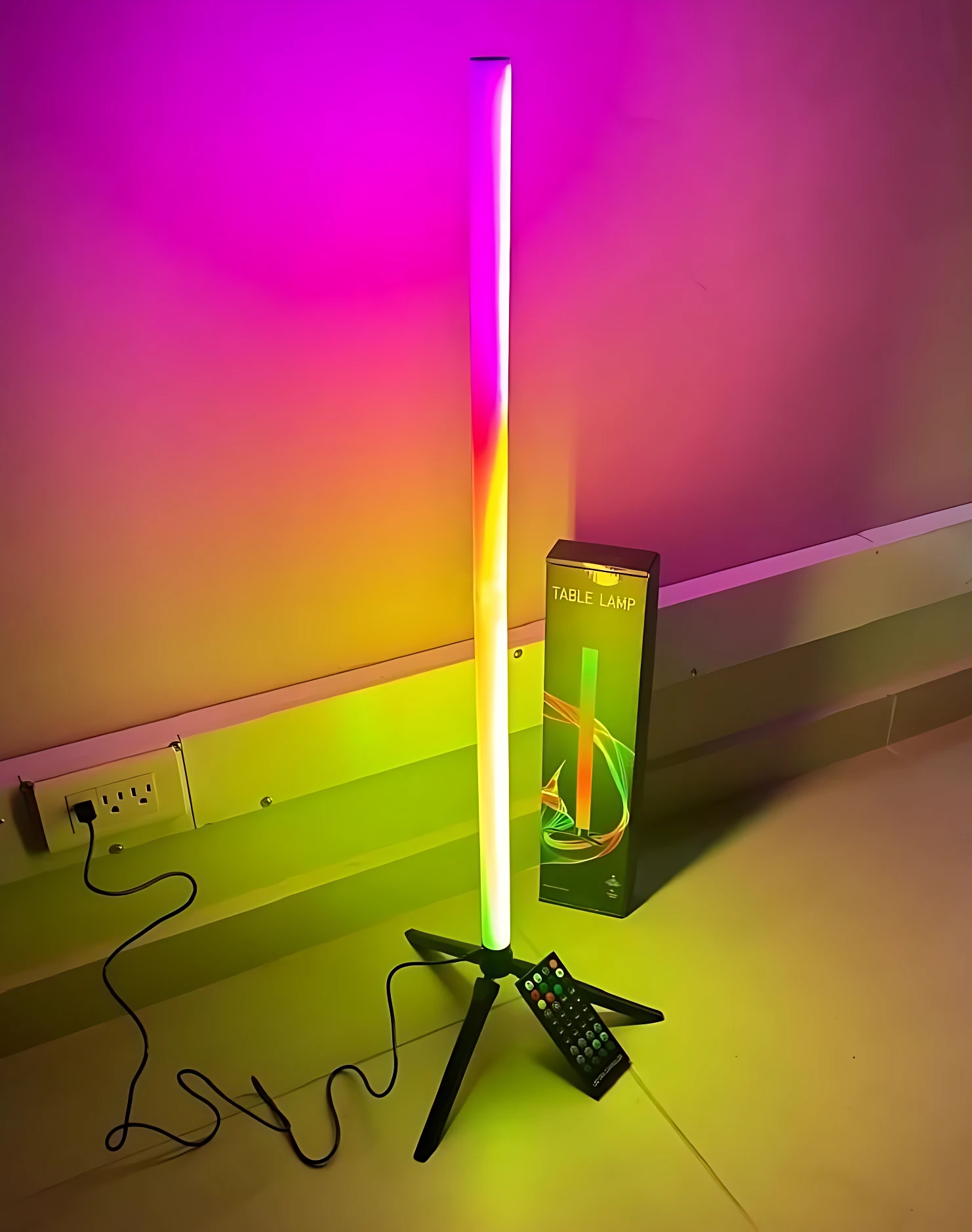 Tubo neón RGB vertical de 75 cm con soporte trípode negro, emitiendo luz multicolor en ambiente interior junto a su caja y control remoto.