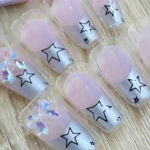 Set de tres uñas postizas con diseño de estrellas negras sobre base plateada y detalles en rosa pastel, presentadas en empaque individual.