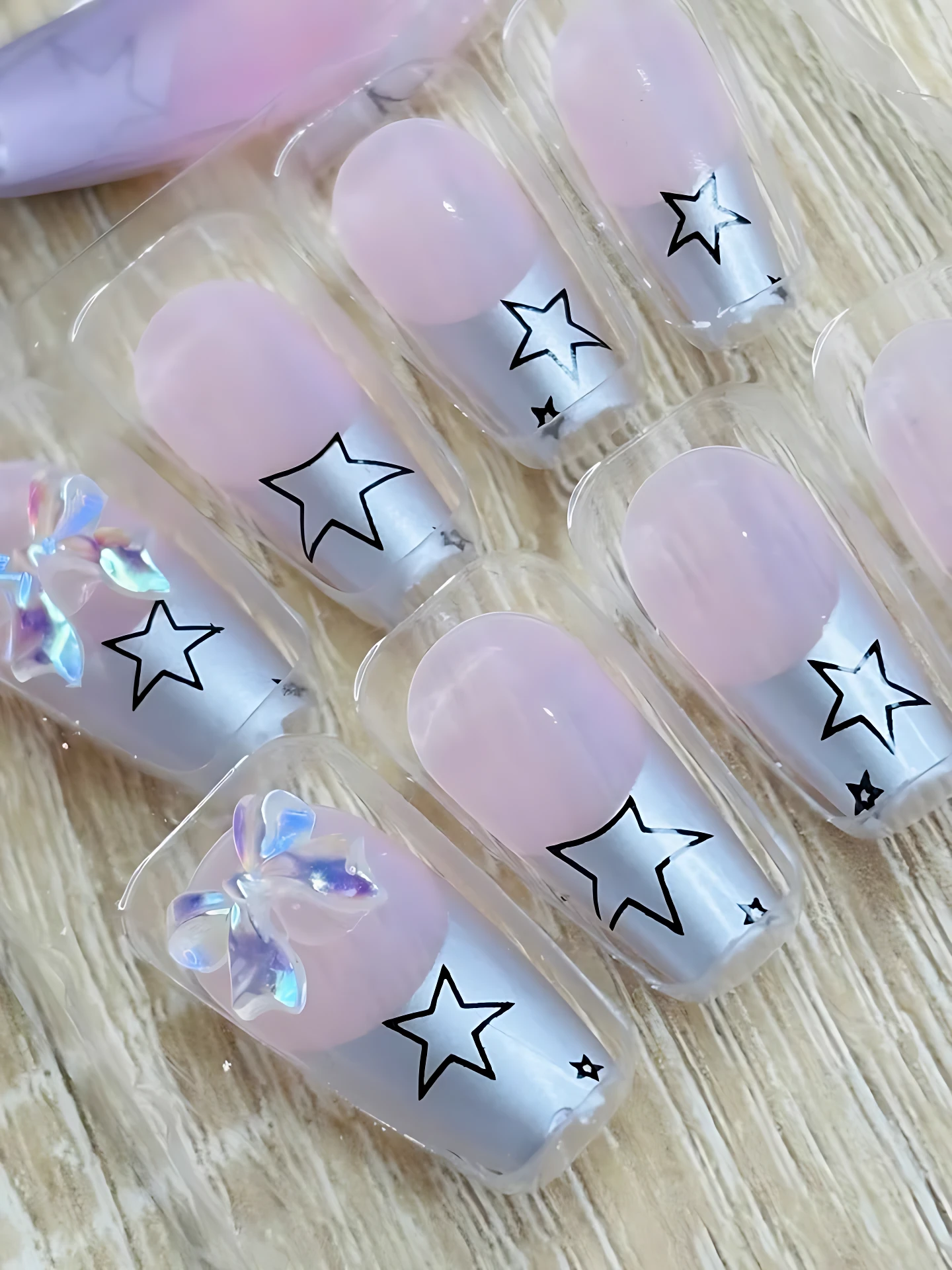Set de tres uñas postizas con diseño de estrellas negras sobre base plateada y detalles en rosa pastel, presentadas en empaque individual.