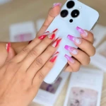Manos con uñas postizas largas en tonos rosa y rojo con detalles blancos, sosteniendo un teléfono blanco sobre fondo con cajas de producto.