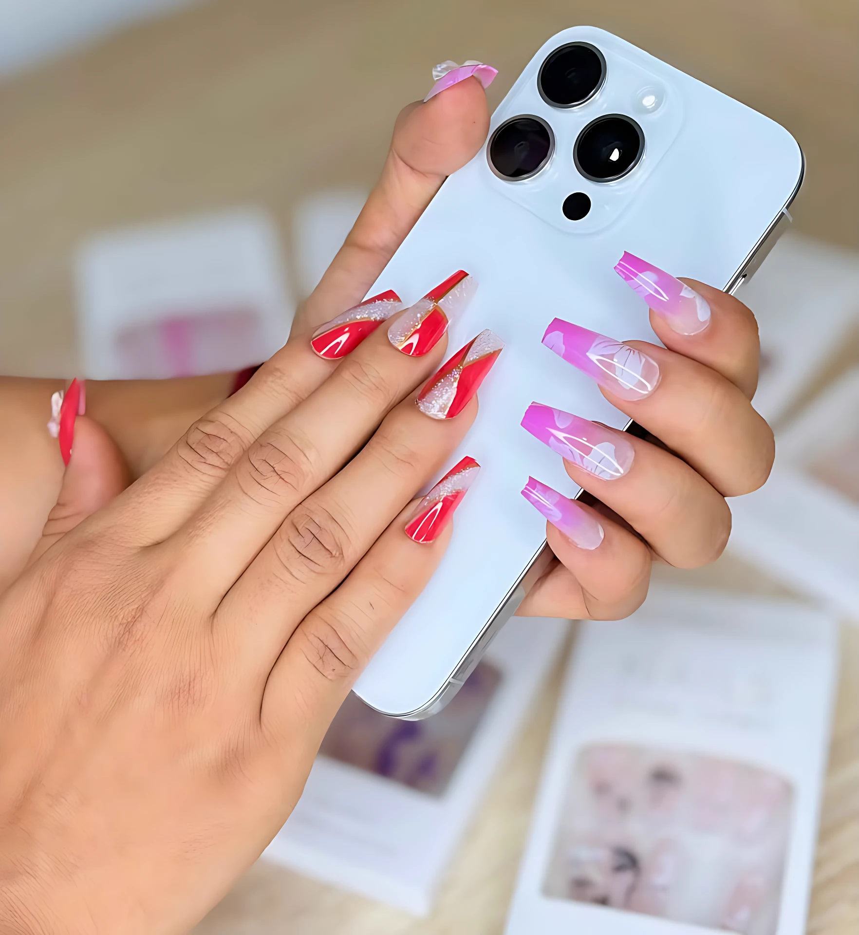 Manos con uñas postizas largas en tonos rosa y rojo con detalles blancos, sosteniendo un teléfono blanco sobre fondo con cajas de producto.