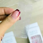 Mano sosteniendo una tira transparente con uñas postizas de forma ovalada, color nude con punta roja, sobre cajas del producto.