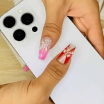 Mano sosteniendo teléfono blanco, mostrando uñas postizas largas con diseños de flores en tonos rosa y rojo sobre base blanca.