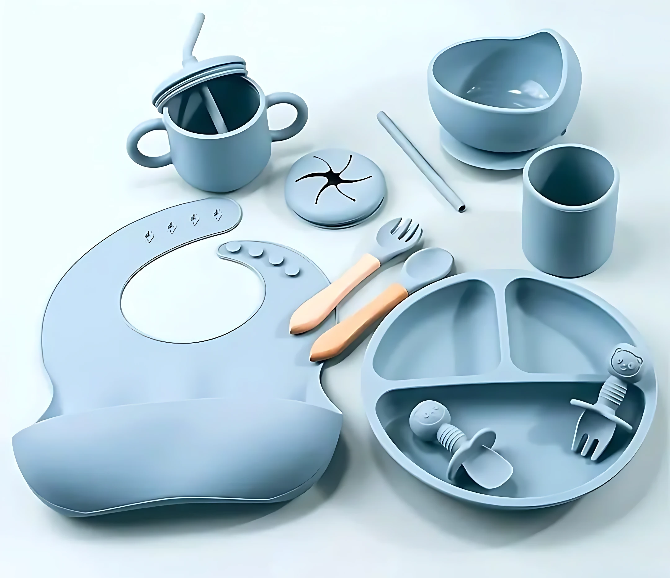 Set de utensilios de bebé en silicona azul claro, con plato dividido, biberón, taza, cubiertos y babero, dispuestos sobre fondo blanco.