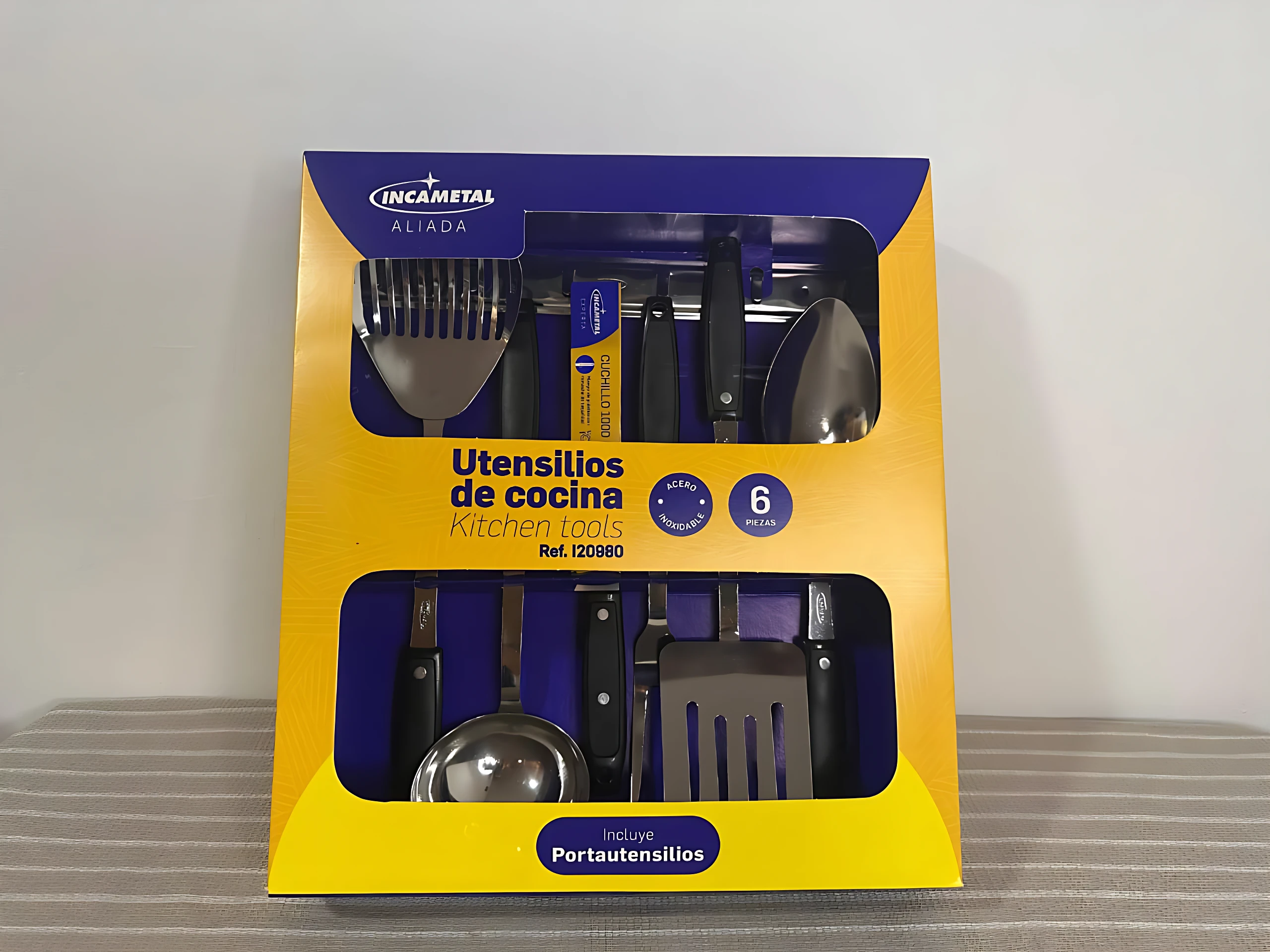 Pack de seis utensilios de cocina en acero inoxidable, con mango negro y cabezales metálicos, presentados en caja amarilla y azul con ventana transparente. Incluye espátula, cuchara, tenedor, espátula de silicona, colador y espátula plana.