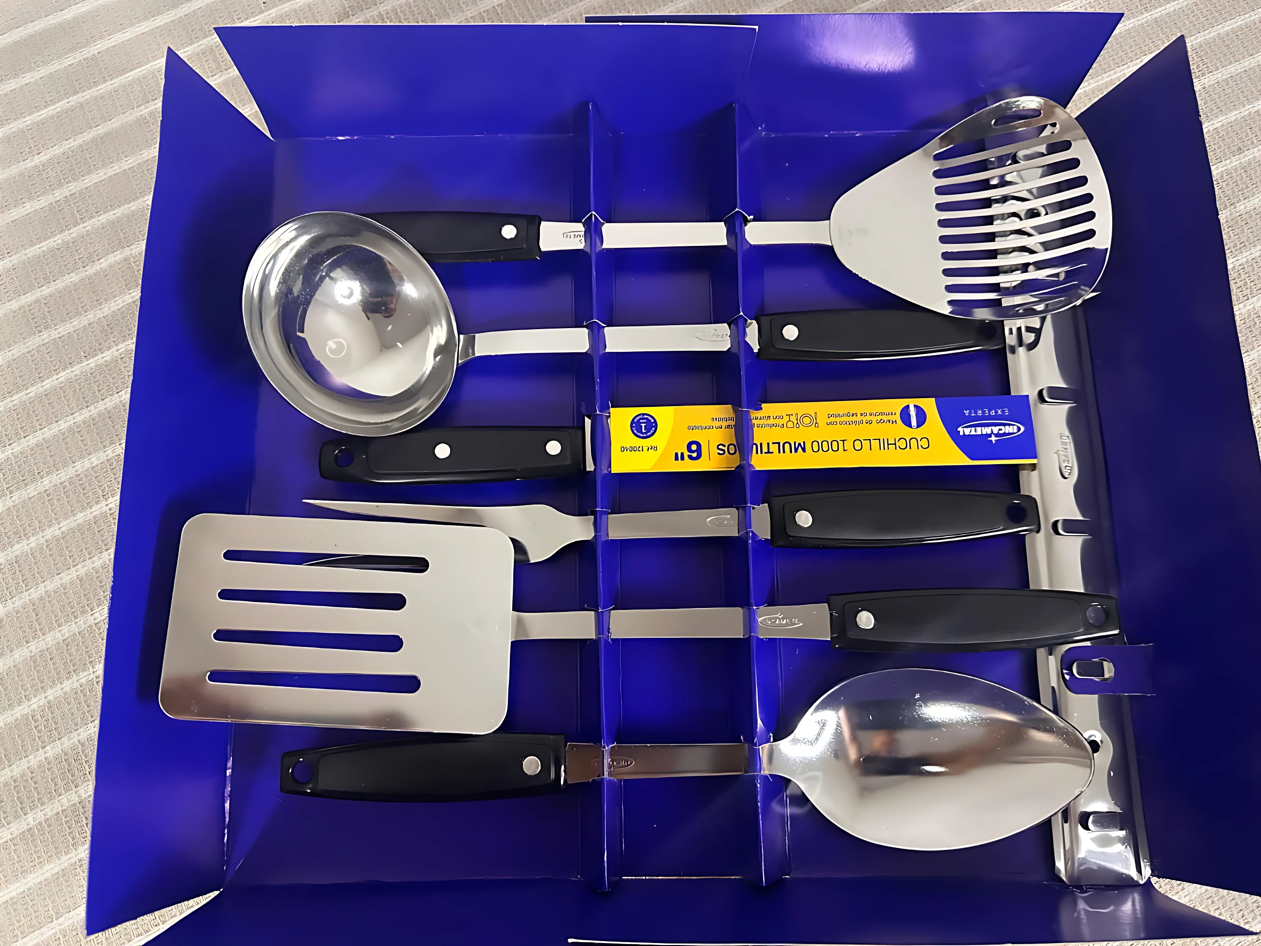 Set de seis utensilios de cocina en acero inoxidable, dispuestos en caja azul con compartimentos individuales, incluye cuchara de servir, espátula, colador, tenedor de parrilla y dos cucharas.