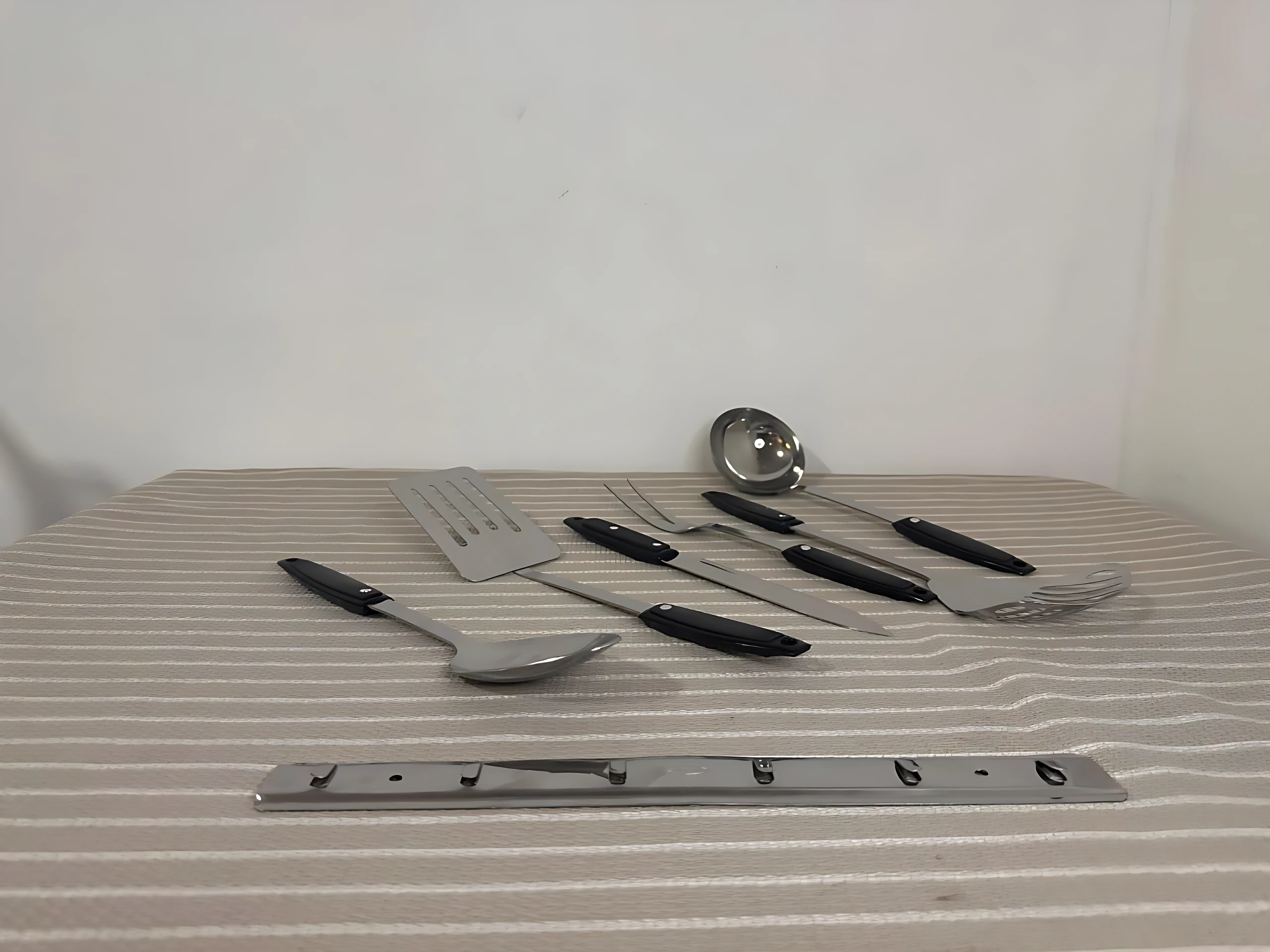 Set de seis utensilios de cocina en acero inoxidable con mangos negros, dispuestos sobre tela a rayas, junto con soporte metálico para colgar.