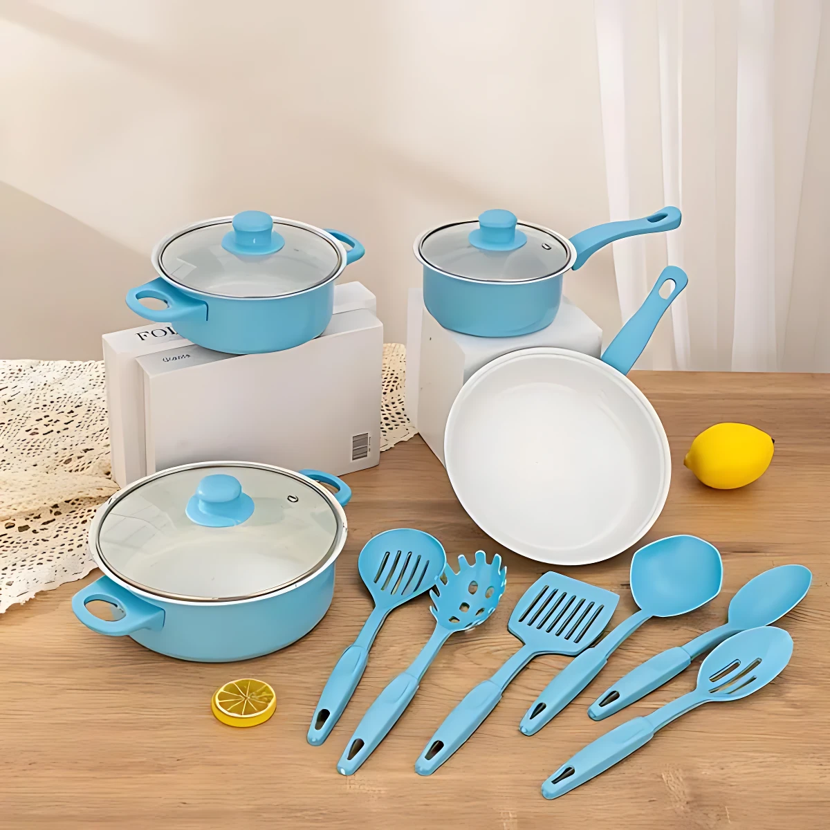 Set de cocina azul con ollas, sartén y utensilios de silicona sobre mesa de madera, junto a limones y cajas de presentación.